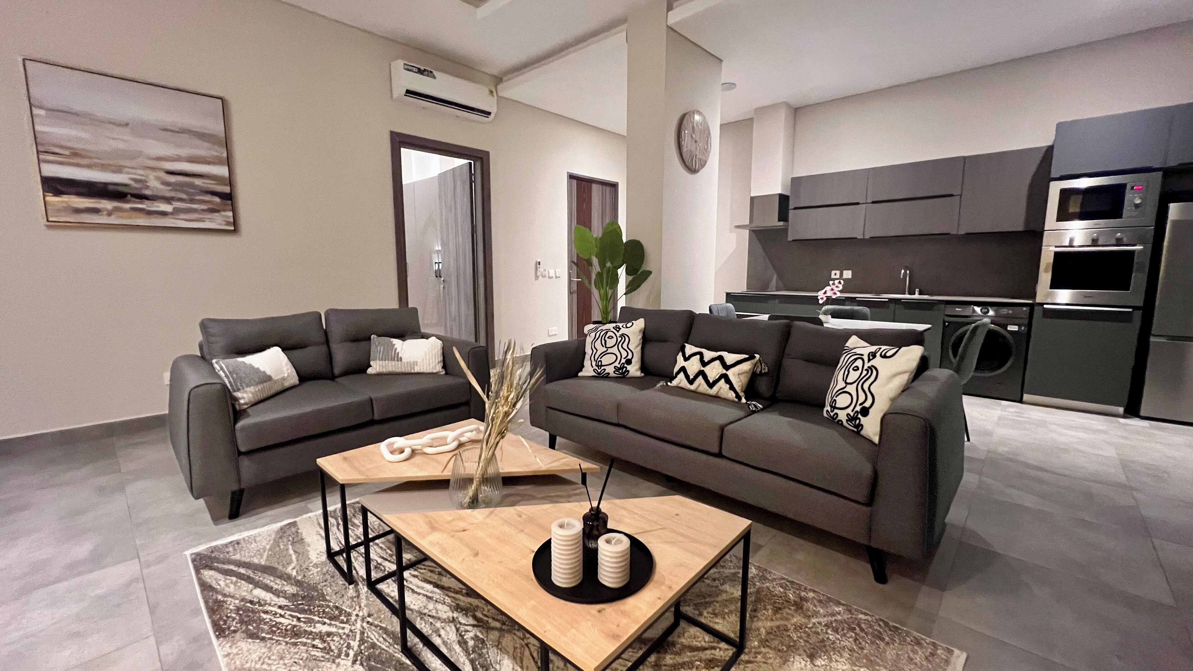 A modern living room with a grey sofa set, a wooden coffee table, and a kitchen area with dark cabinetry and appliances.