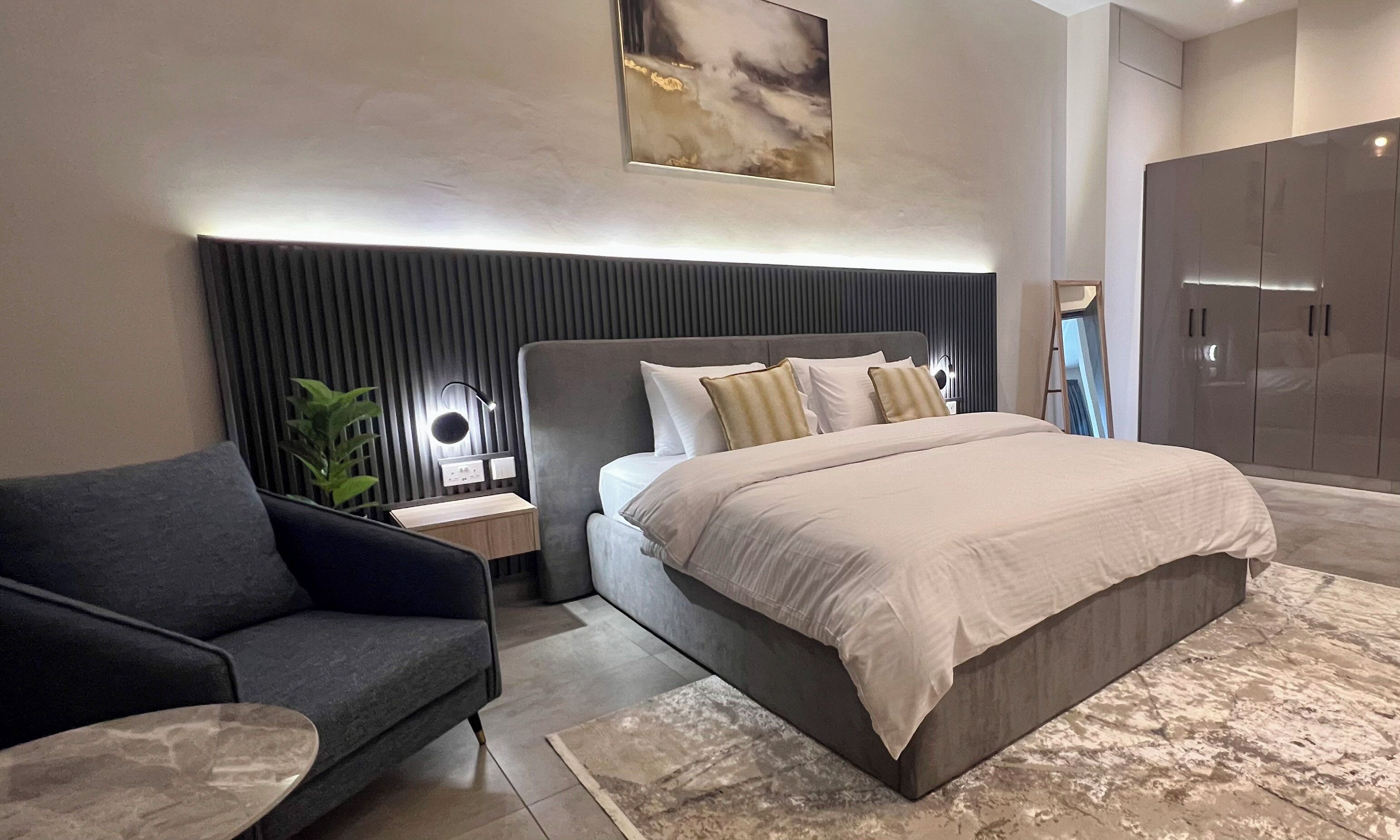 Un dormitorio moderno con una cama grande, un sofá gris, una mesita de noche, una lámpara de pared y un armario.