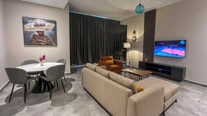 A modern living room with a beige sofa, a dining table with chairs, a TV mounted on the wall, and a large window with curtains. - Anthonian Suites (Accra)