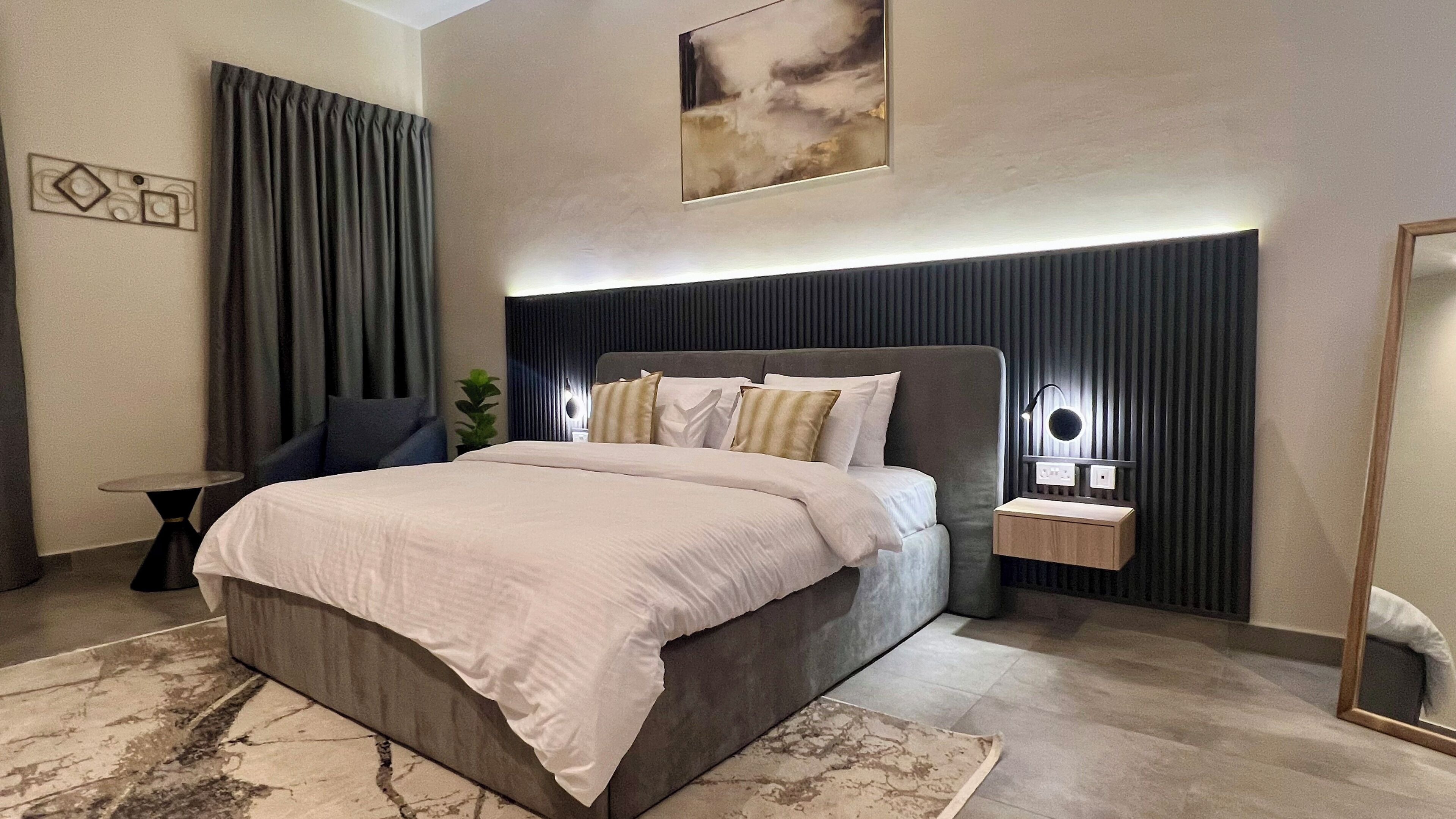 Une chambre moderne avec un grand lit, une table de chevet avec une lampe, un miroir et une œuvre d’art accrochée au mur.