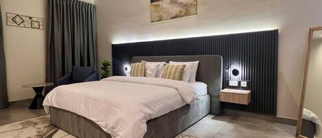 A modern bedroom with a large bed, a bedside table with a lamp, a mirror, and a wall-mounted artwork.