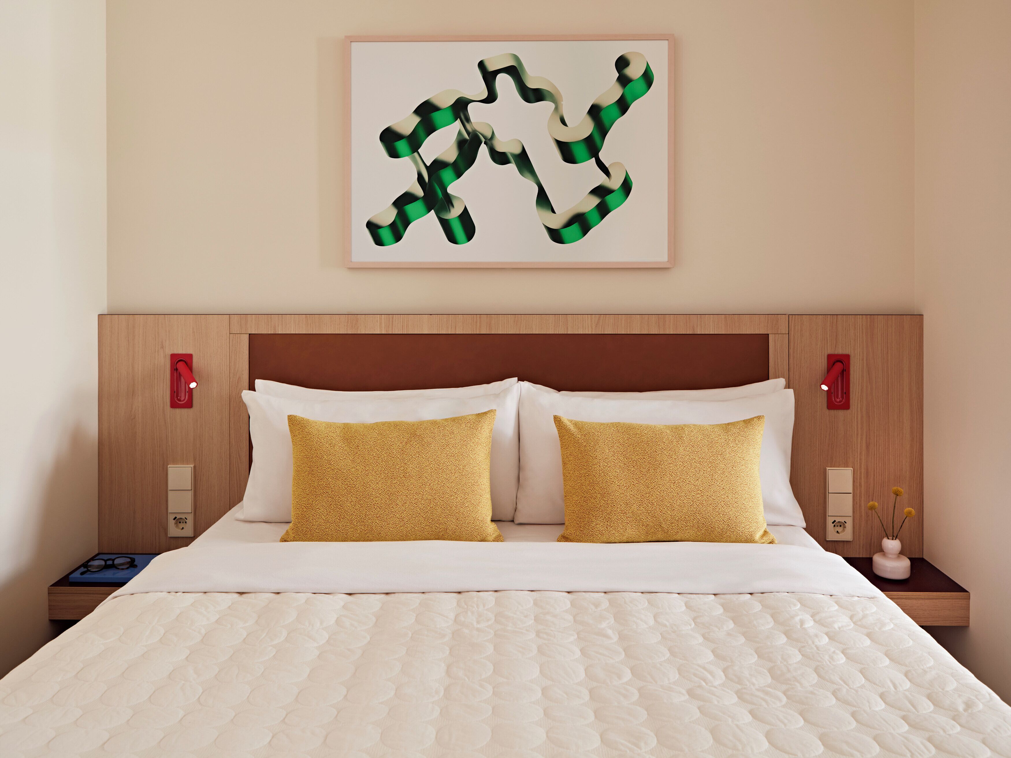 Una cama bien hecha con colcha blanca y dos almohadas doradas, un cabecero de madera y una obra de arte abstracta enmarcada en la pared.