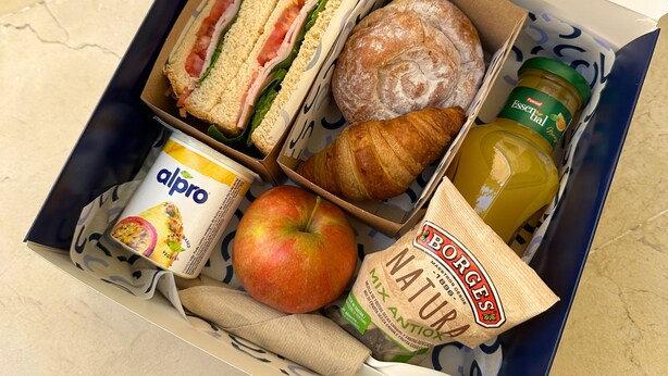 Una fiambrera con sándwiches, un croissant, una manzana, una botella de jugo, un envase de yogur y una bolsa de pan.
