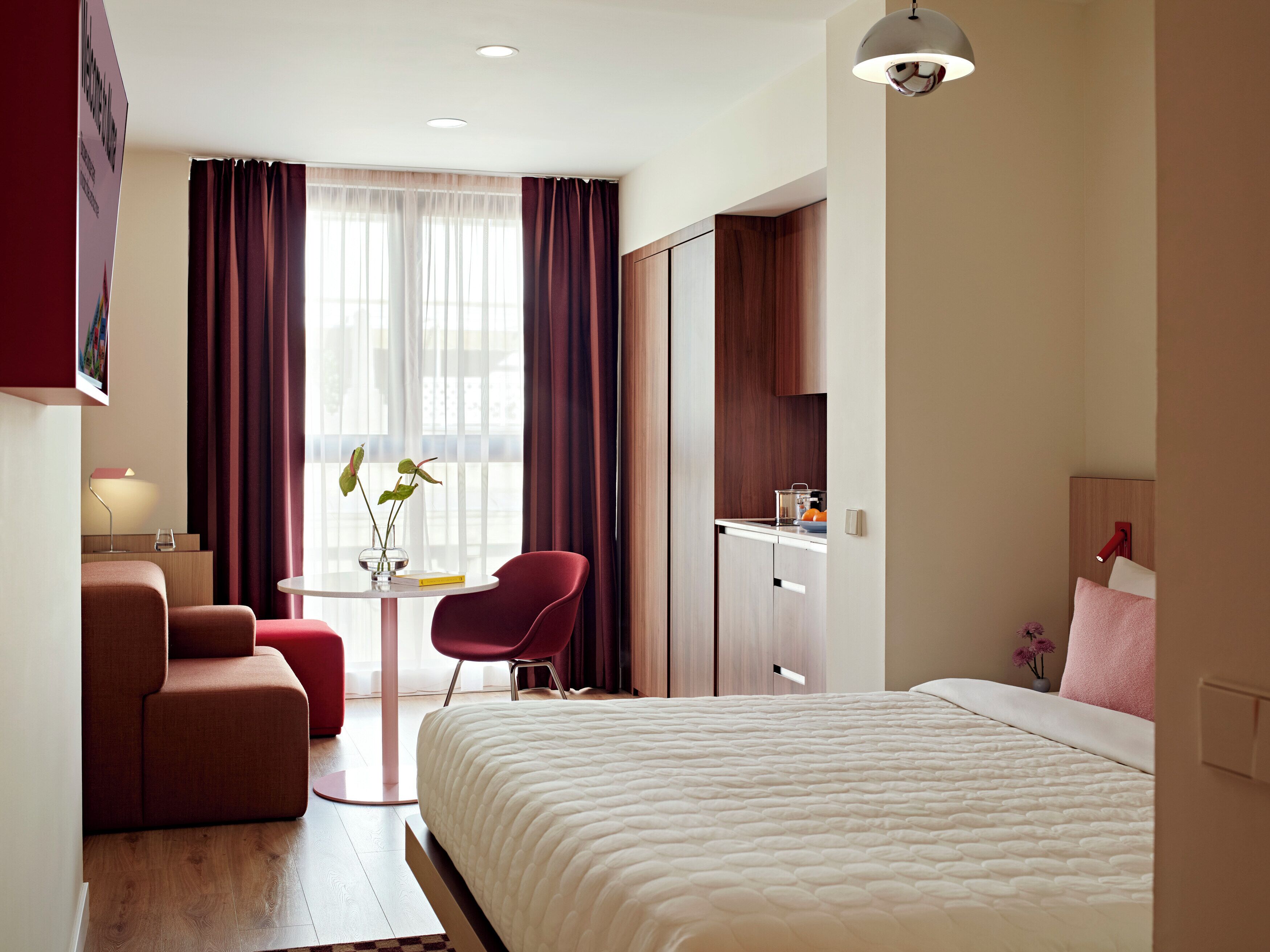 A hotel room with a bed, a desk, a chair, a small table with a vase, and a view of the outside through the window.