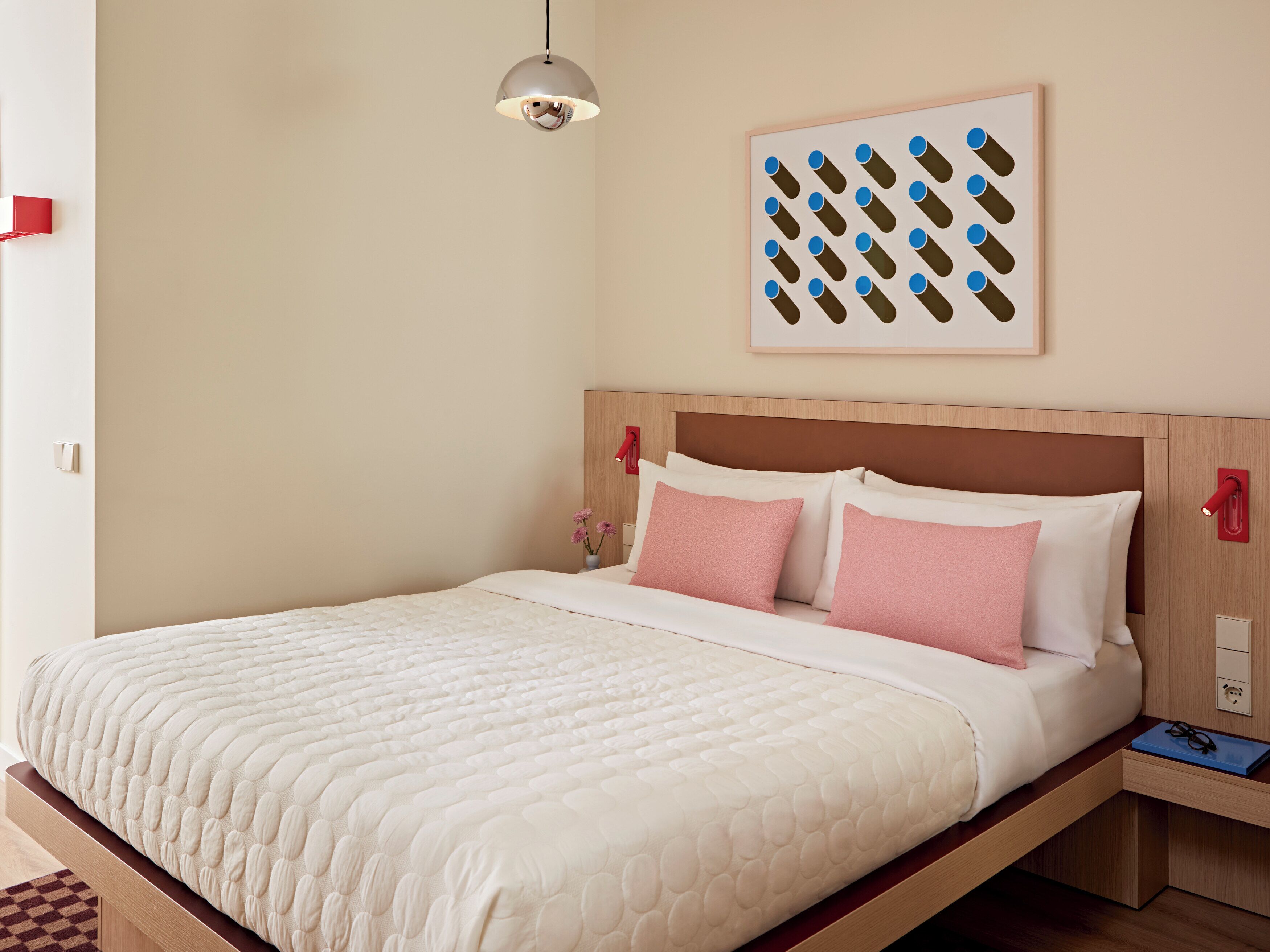 A neatly made bed with a white quilt and pink pillows, a wooden headboard, a wall-mounted light fixture, and a framed picture above the bed.