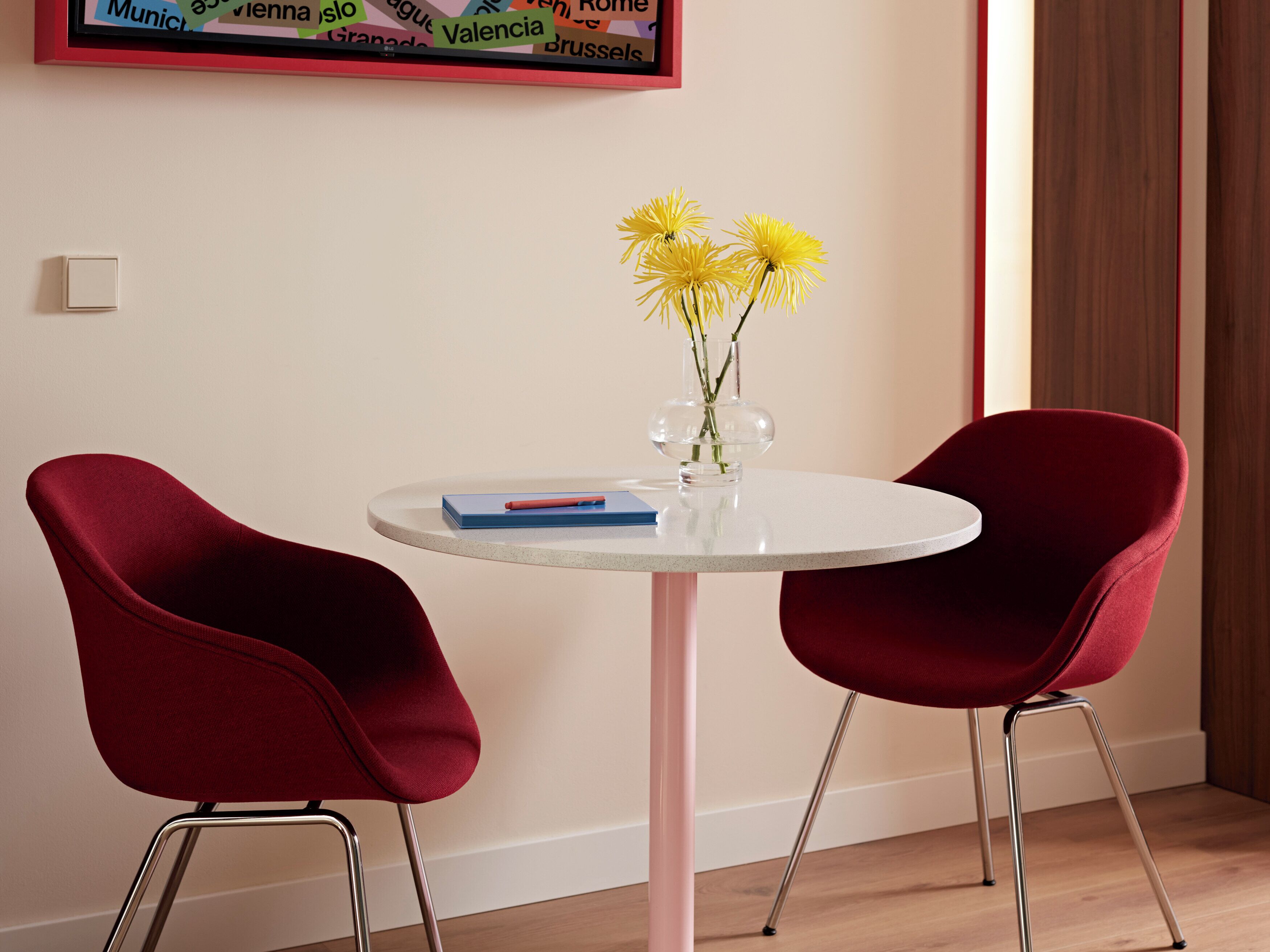 A room with a round table, two red chairs, a vase with yellow flowers, a blue book, and a framed map on the wall.