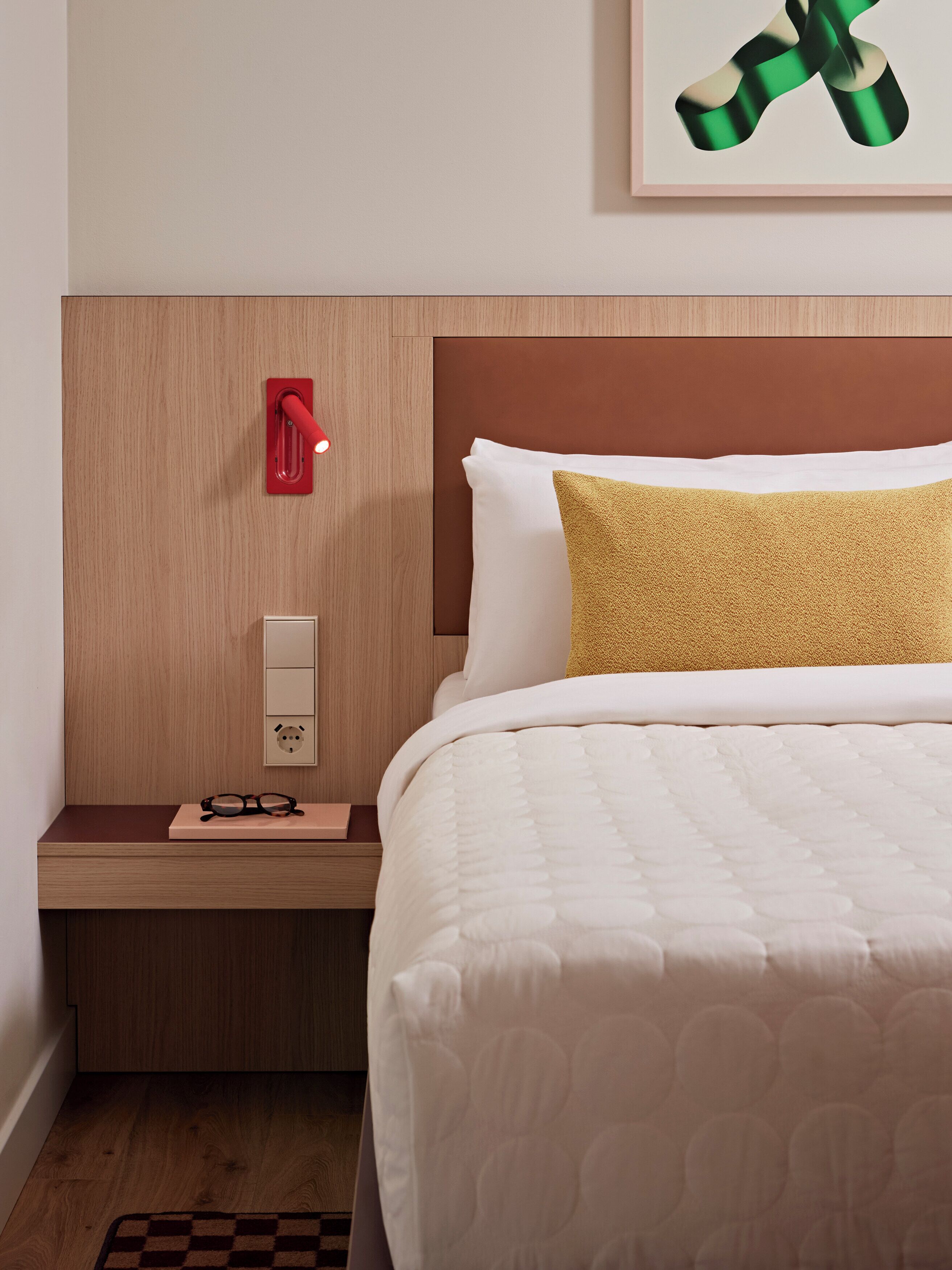 A neatly made bed with a white comforter and a gold accent pillow, a bedside table with a pair of glasses, a red wall-mounted lamp, and a framed artwork above the bed.