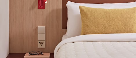 A neatly made bed with a white comforter and a gold accent pillow, a bedside table with a pair of glasses, a red wall-mounted lamp, and a framed artwork above the bed.