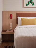 A neatly made bed with a white comforter and a gold accent pillow, a bedside table with a pair of glasses, a red wall-mounted lamp, and a framed artwork above the bed.
