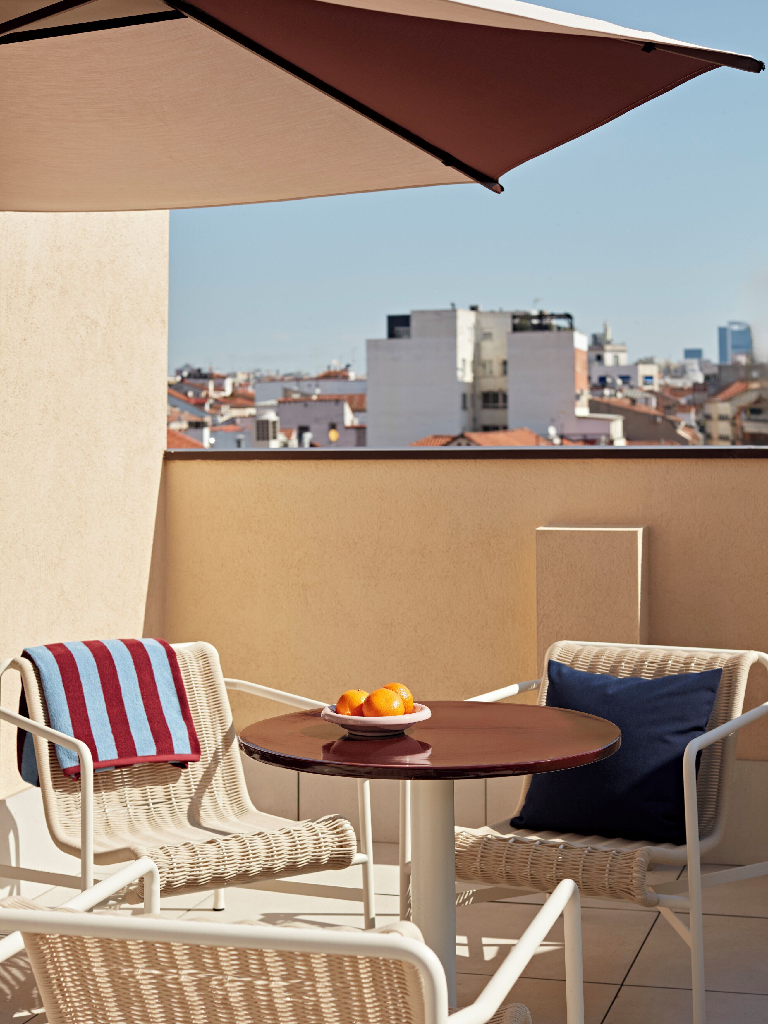 A balcony with wicker chairs, a table with a bowl of fruit, and a view of a cityscape.