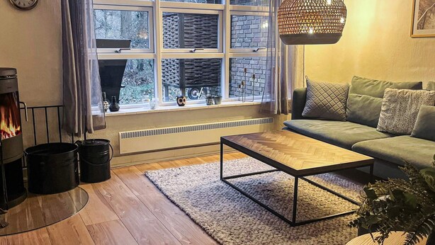 Ein gemütliches Wohnzimmer mit Kamin, einem grünen Sofa, einem Holztisch und einem großen Fenster mit Blick auf Bäume.