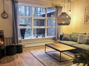 Ein gemütliches Wohnzimmer mit Kamin, einem grünen Sofa, einem Holztisch und einem großen Fenster mit Blick auf Bäume.