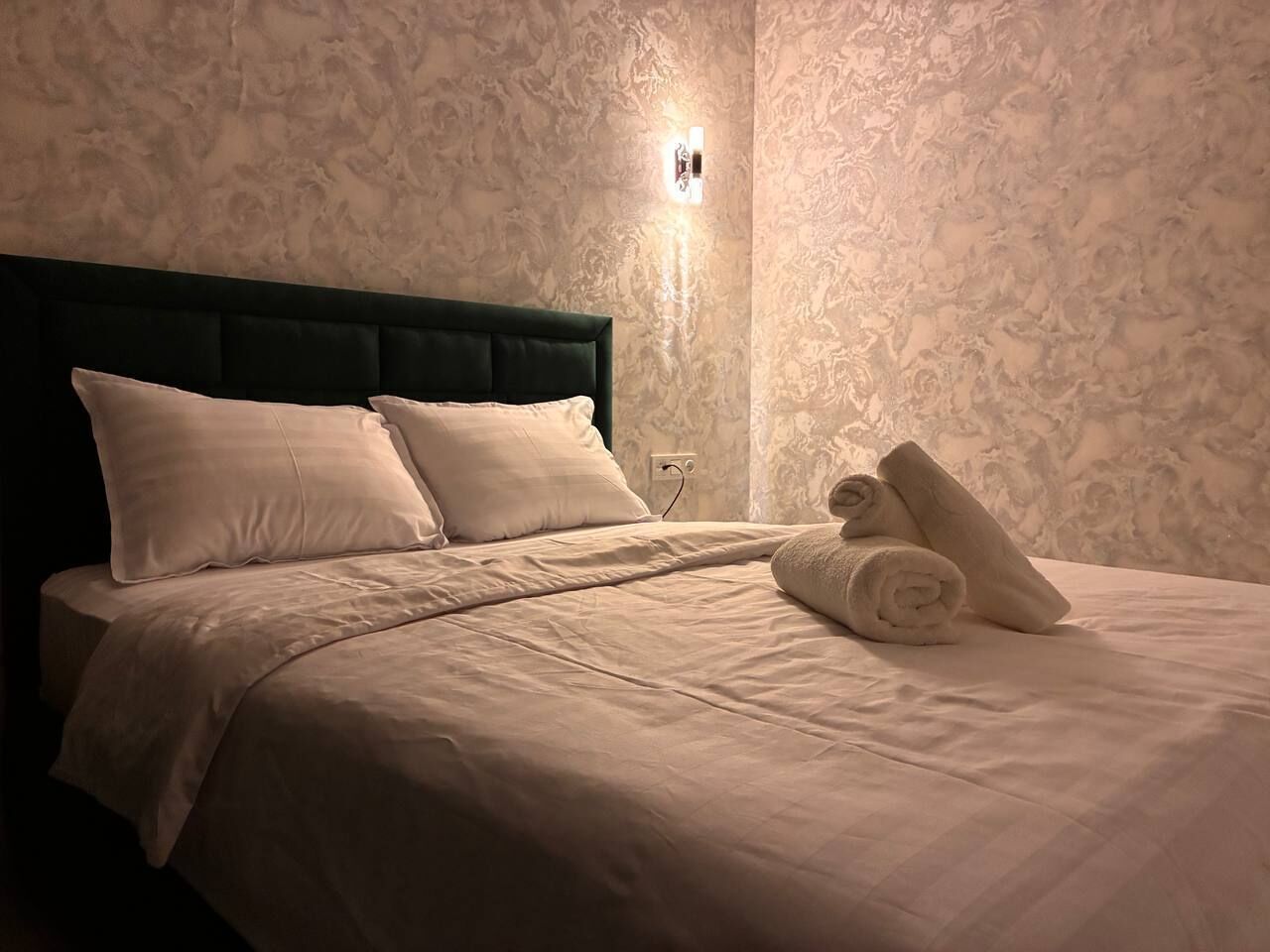 Una cama bien hecha con un cabecero oscuro, una lámpara fijada a la pared y toallas enrolladas sobre la cama.