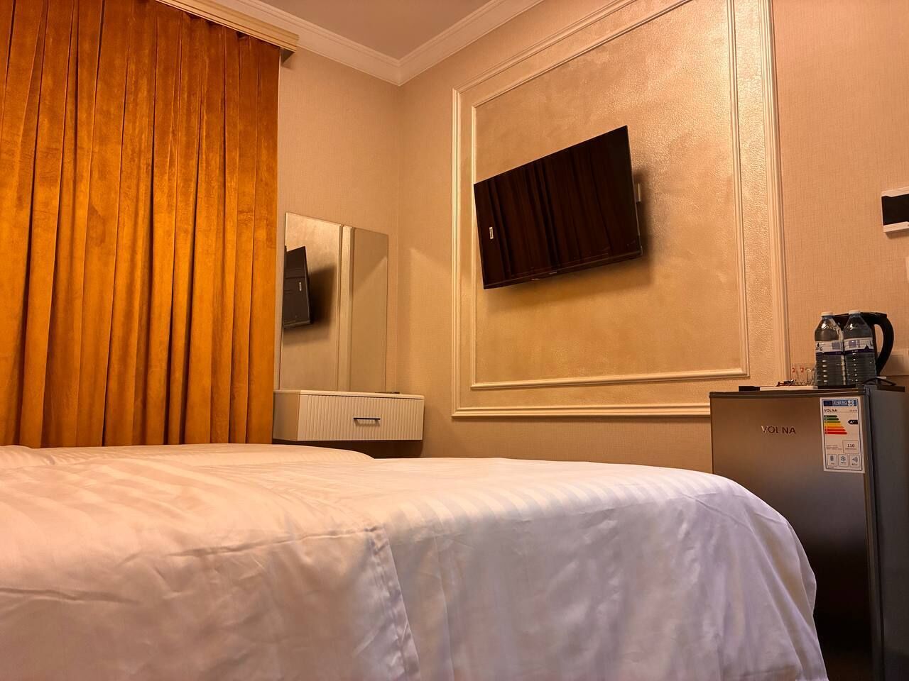 Quarto de hotel com cama, minibar, televisão fixada na parede e janela com cortinas.
