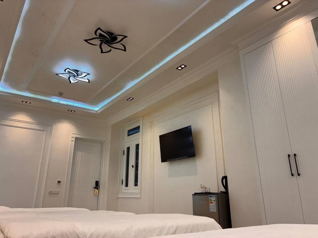 A hotel room with two beds, a flat-screen TV, a minibar, and a ceiling with recessed lighting and decorative elements.
