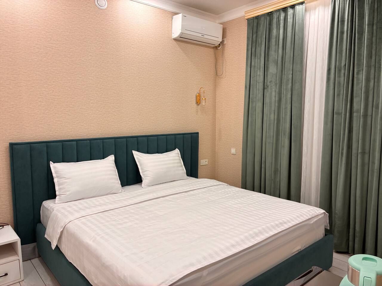 A neatly made bed with a green headboard, a white pillow, and a white bedspread. There is a small bedside table with a lamp, and a green curtain partially covering a window.