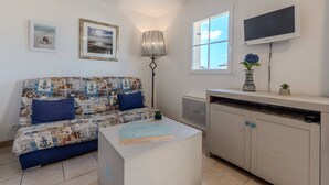 A living room with a patterned sofa, a small table with books, a lamp, a TV, and a cabinet. - Peaceful Coastal Stay With Access to Pool (Saint-Nic)