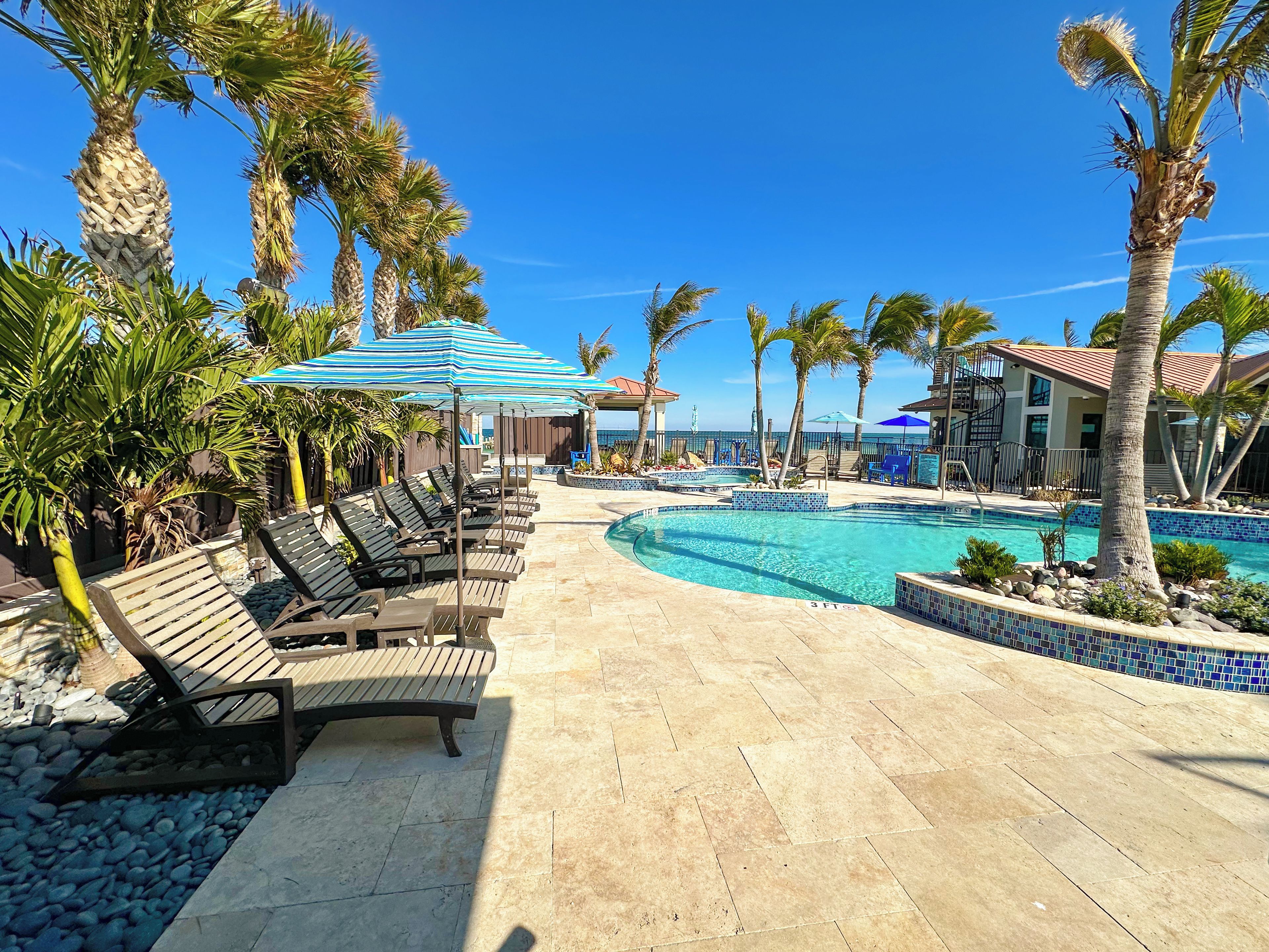 A poolside area with lounge chairs and umbrellas, a clear blue sky, and a view of the ocean.