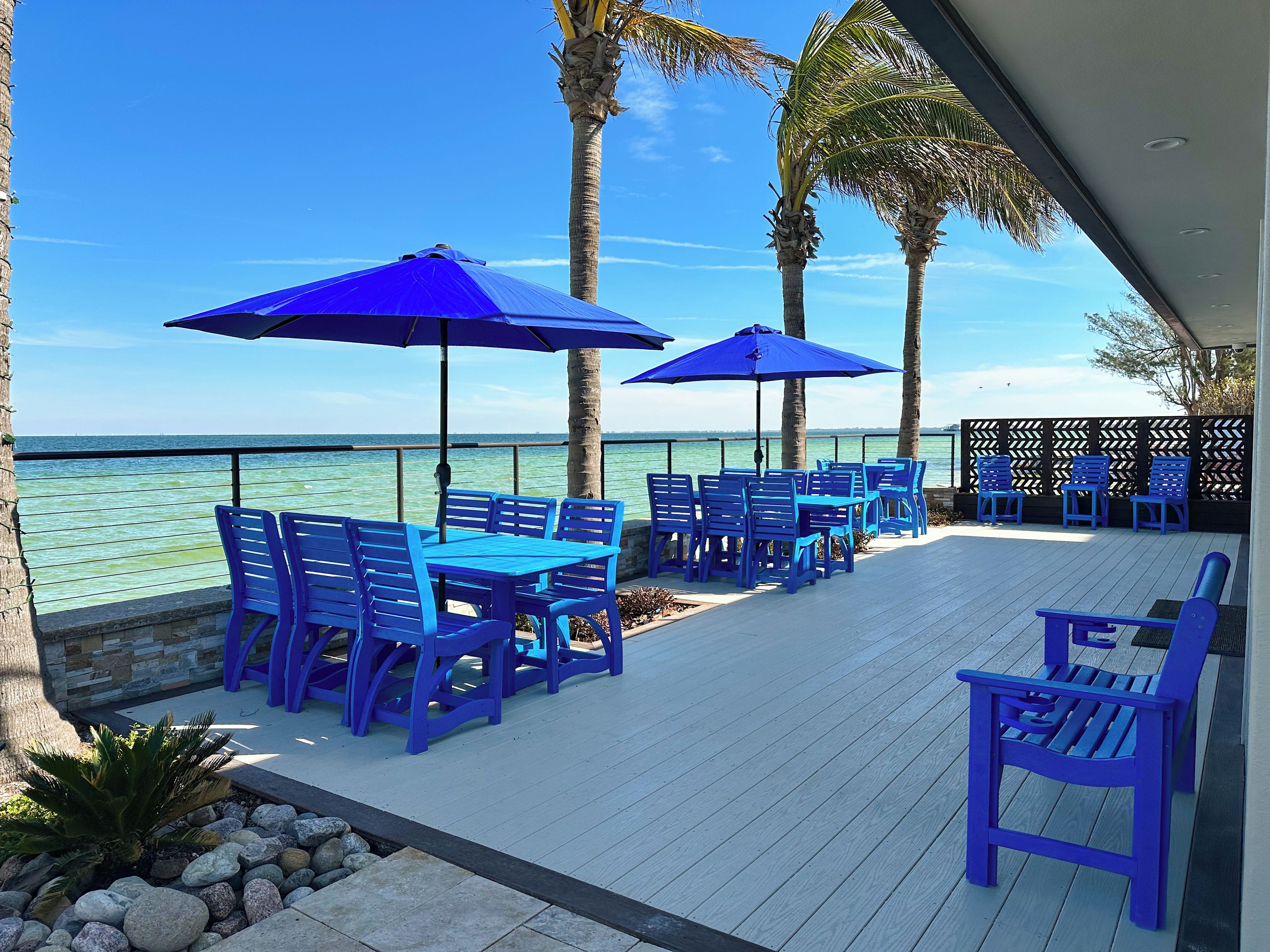A deck with blue chairs and tables, palm trees, and a view of the ocean.