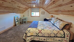 A cozy attic bedroom with a wooden ceiling, a bed with a colorful quilt, a desk with a chair, and a window with a view. - Beautiful, newly renovated 4-bedroom, 2 bath farmhouse in the Petoskey area (Petoskey)