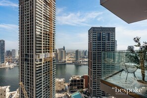 A high-rise building with a construction crane, a glass balcony with a chair, and a view of a cityscape and water body. - Luxury 2BR in Peninsula One by Bespoke (Dubai)