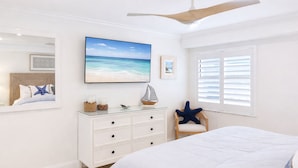 Un dormitorio con cama, cómoda, silla, ventilador de techo y ventana con persianas, además de un cuadro con temática de playa en la pared.