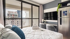 Moderni hotellihuone, jossa on suuri sänky, oleskelualue, televisio ja parveke, jolta on näkymä kaupunkiin.