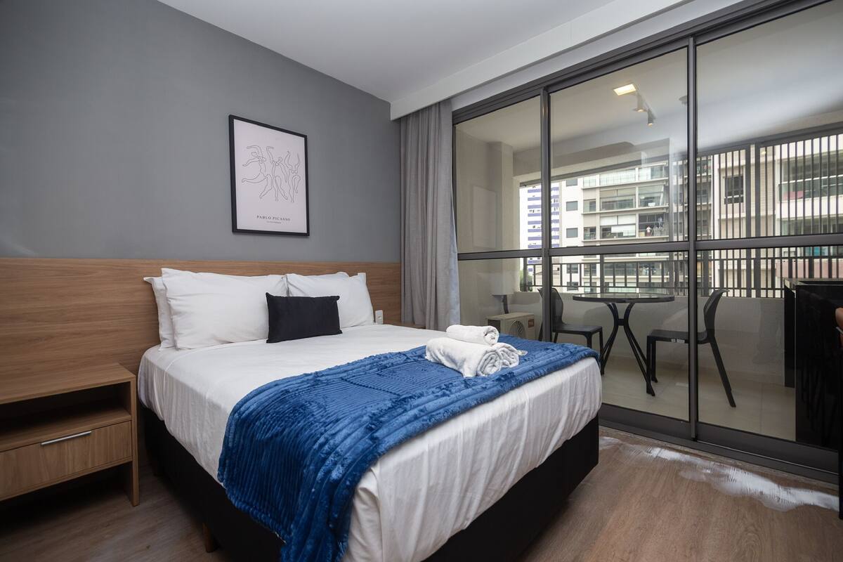 Une chambre d’hôtel avec un grand lit, une table de chevet en bois, une œuvre d’art encadrée au mur et un balcon offrant une vue sur les bâtiments.