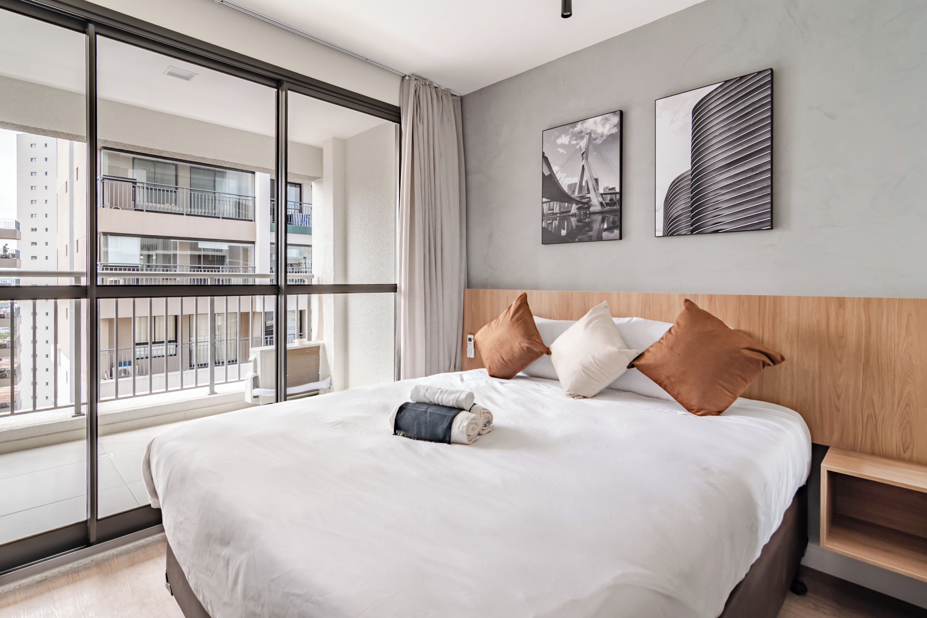 Habitación de hotel moderna con una cama grande, cabecera de madera y vistas a un paisaje urbano a través de ventanas de techo a suelo.