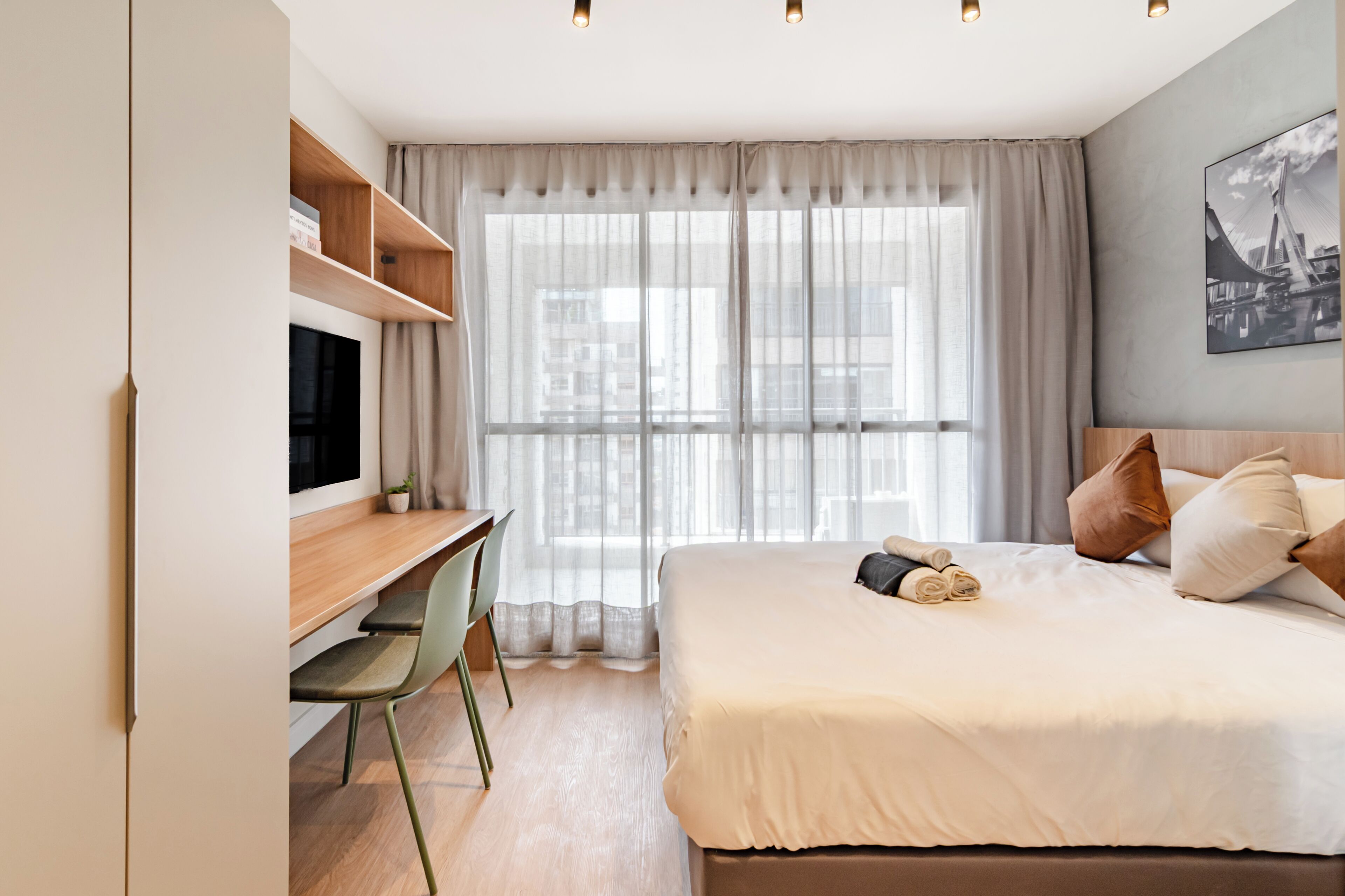 A modern hotel room with a large bed, a desk with a chair, a TV, and a window with sheer curtains.