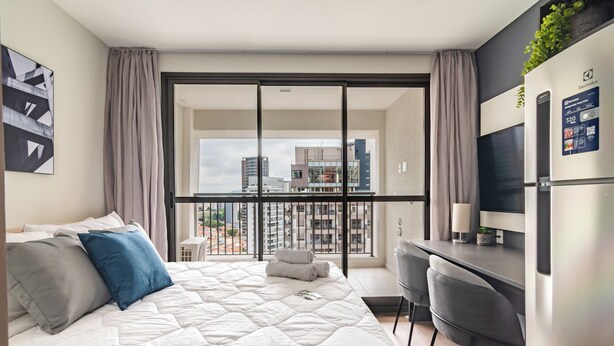 Moderni hotellihuone, jossa on sänky, työpöytä, tuoli, jääkaappi ja suuri ikkuna, josta avautuu näkymä kaupunkiin.