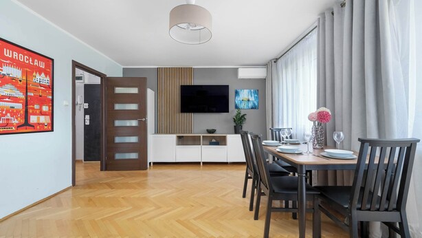A modern dining area with a wooden floor, a dining table with chairs, a TV mounted on the wall, and a window with curtains. - Spacious Apartment in Wroclaw | Air Conditioning and Balcony (Wrocław)