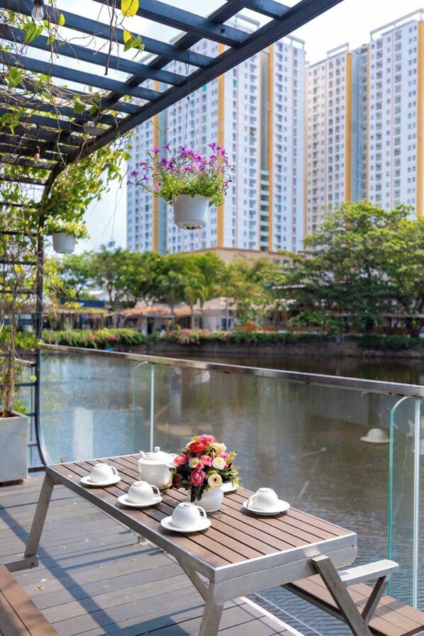 Una mesa de madera con teteras blancas y un ramo de flores en un balcón con vistas a un río, con edificios altos al fondo.