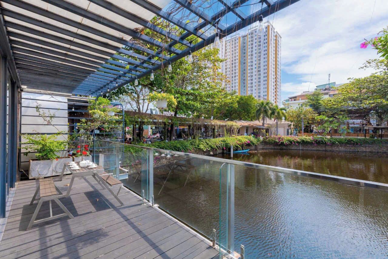A modern outdoor area with a glass railing overlooking a water body, surrounded by greenery and urban buildings.