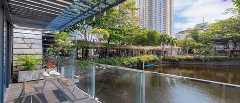 A modern outdoor area with a glass railing overlooking a water body, surrounded by greenery and urban buildings.