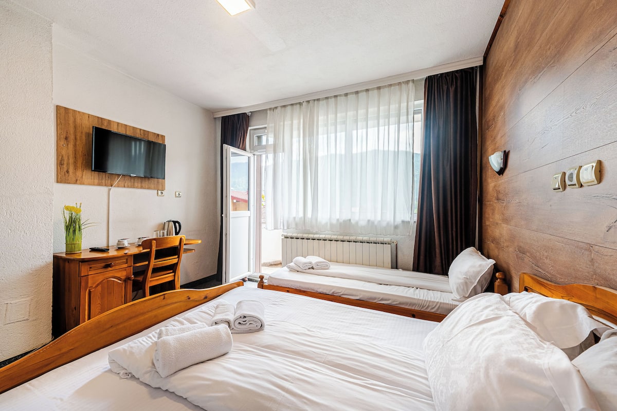 A hotel room with two beds, a wooden desk, a flat-screen TV mounted on the wall, and a window with curtains.