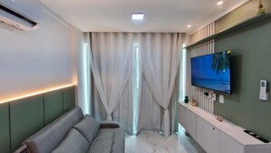 Un salon moderne avec un canapé, un téléviseur fixé au mur et une fenêtre ornée de rideaux légers.