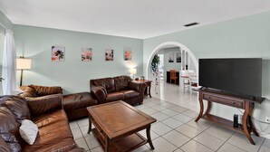 A living room with a brown leather sofa, a wooden coffee table, and a flat-screen TV on a wooden stand. There are framed pictures on the wall and a white archway leading to another room. - Chelsea Way Getaway Bright 4br Family Retreat 4 Bedroom Home (Port Orange)