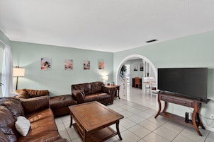 Una sala de estar con un sofá de cuero marrón, una mesa de centro de madera y un televisor de pantalla plana sobre un mueble de madera. Hay cuadros enmarcados en la pared y un arco blanco que conduce a otra habitación.
