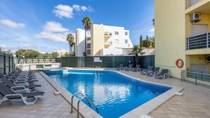 En swimmingpool med liggestole og en redningskrans, omgivet af en bygning med altaner og aircondition-anlæg.