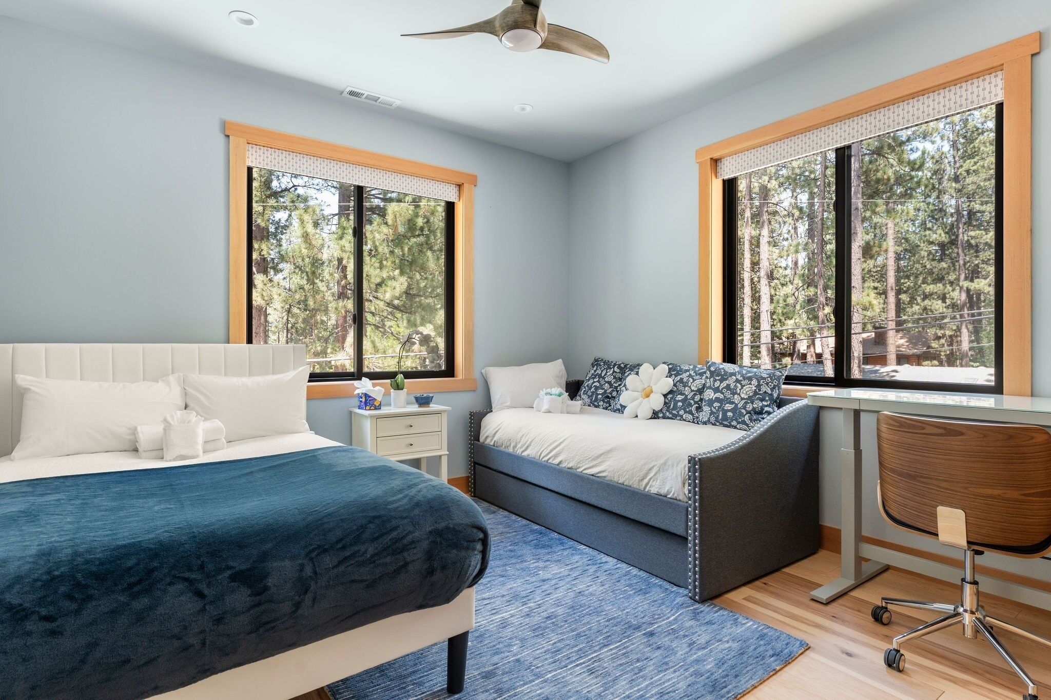 A modern bedroom with two beds, a desk, and large windows offering a forest view.