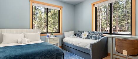 A modern bedroom with two beds, a desk, and large windows offering a forest view.