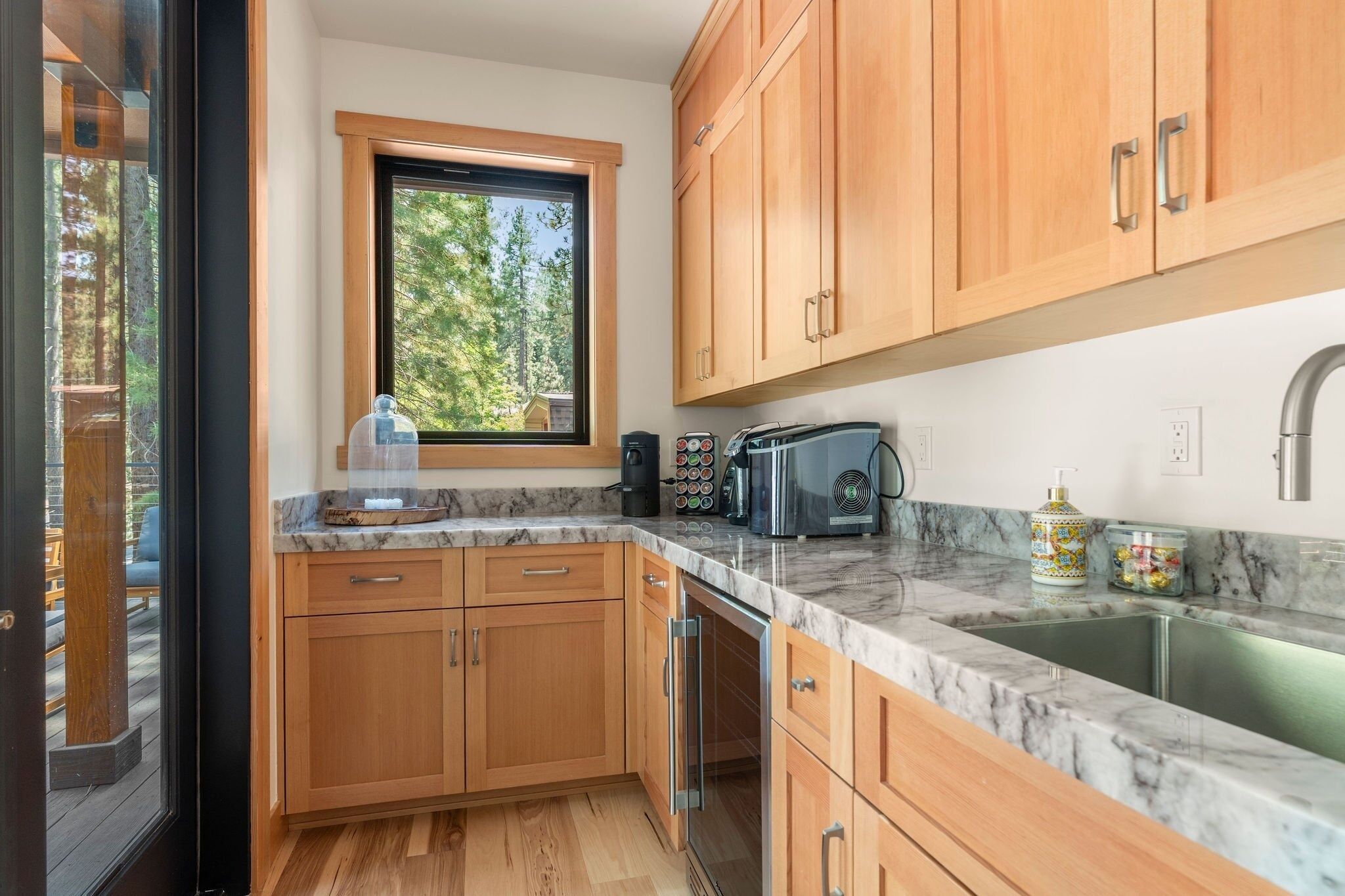 A modern kitchen with wooden cabinets, a marble countertop, a built-in oven, and a window overlooking a forest.