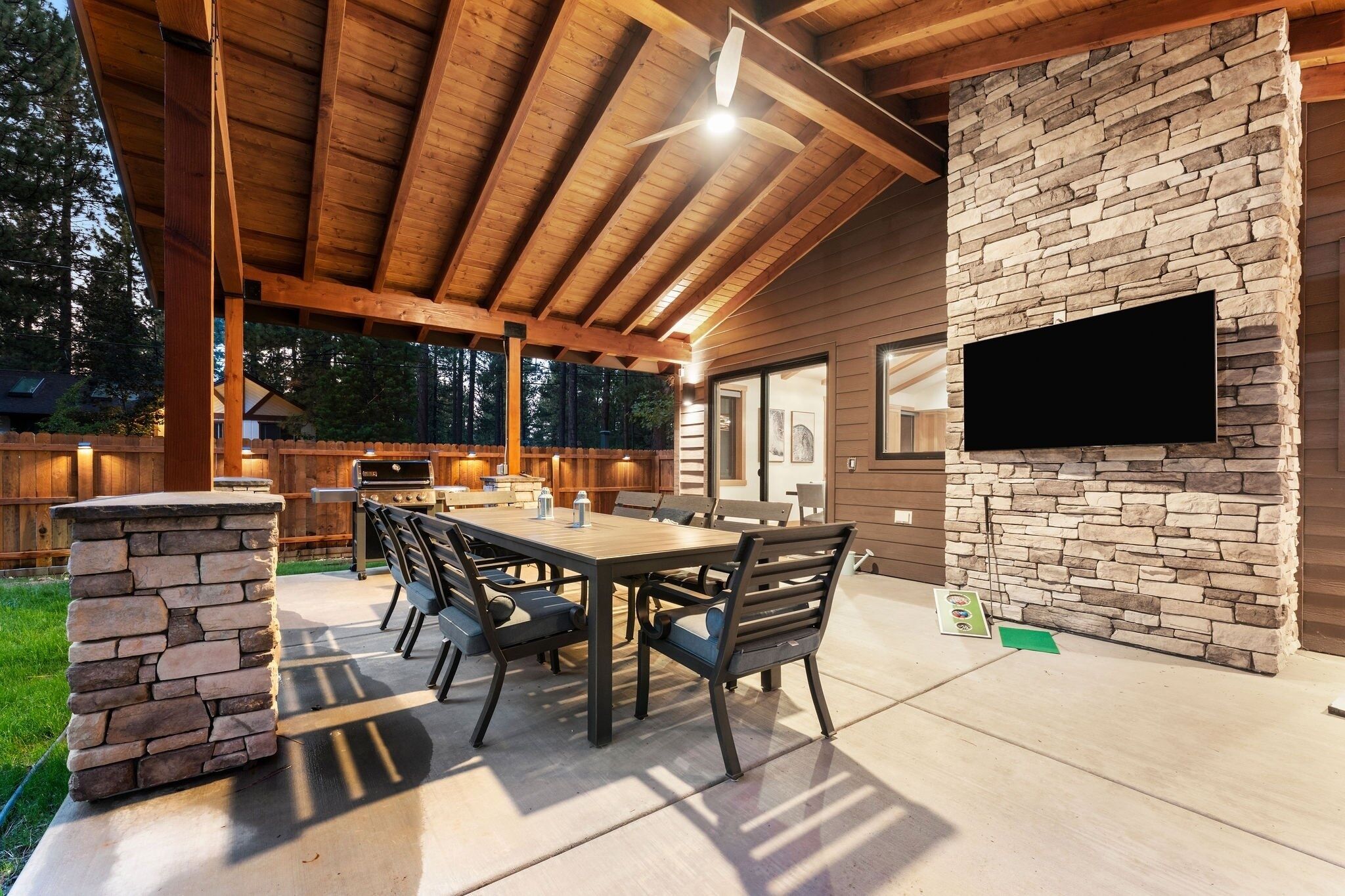 An outdoor patio with a stone fireplace, wooden ceiling beams, and a flat-screen TV mounted on the wall.
