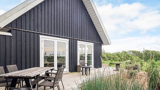 Un bâtiment en bois sombre, doté d’un toit en pente, de grandes fenêtres et d’une terrasse en bois aménagée avec du mobilier d’extérieur.
