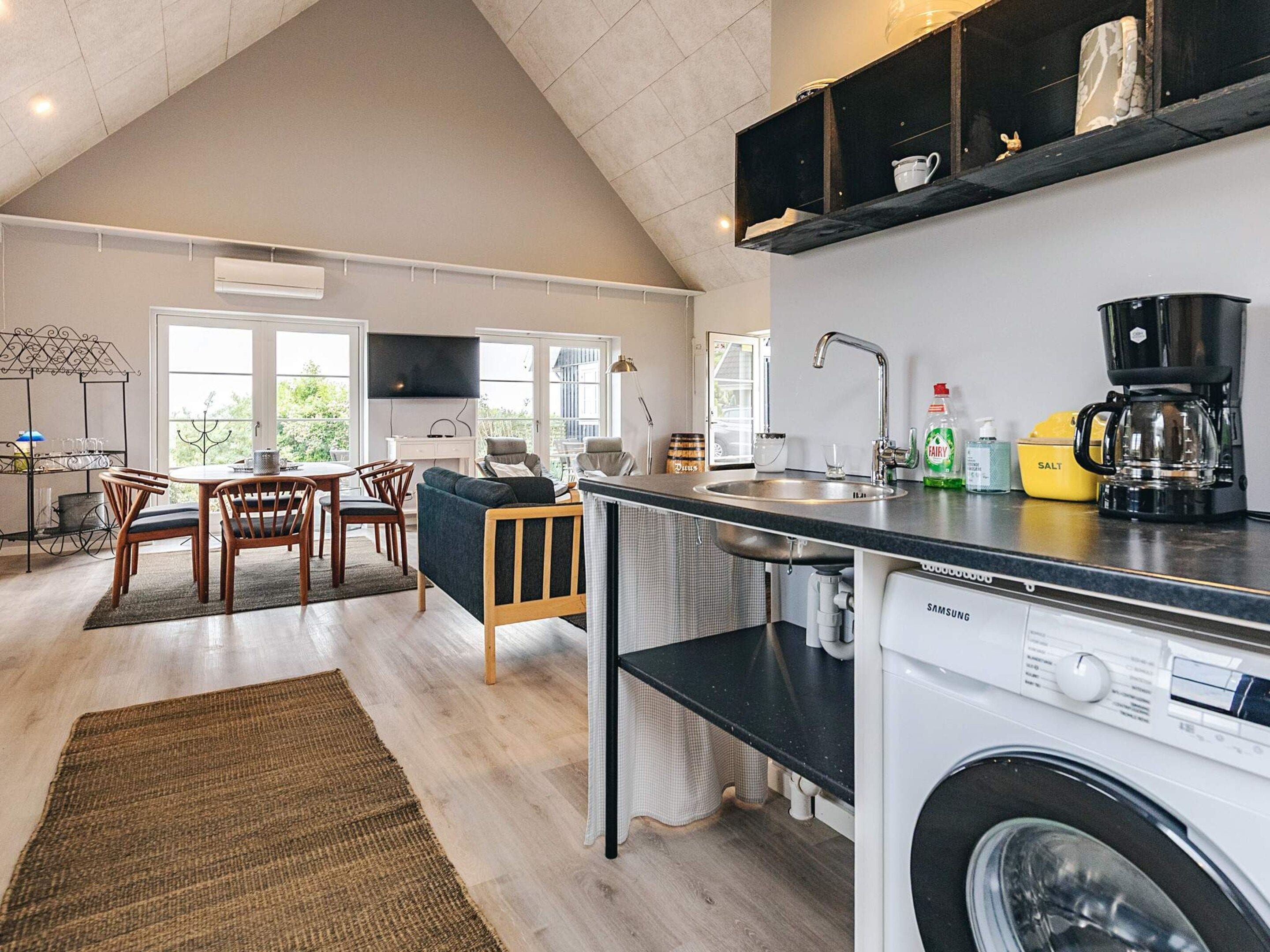 A modern kitchen with a built-in washer and dryer, a dining area with a round table and chairs, and a large window providing natural light.
