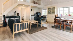 Un vaste espace de vie doté d’un plafond haut, d’un escalier, d’un coin repas avec des meubles en bois et d’une cuisine avec des armoires blanches.