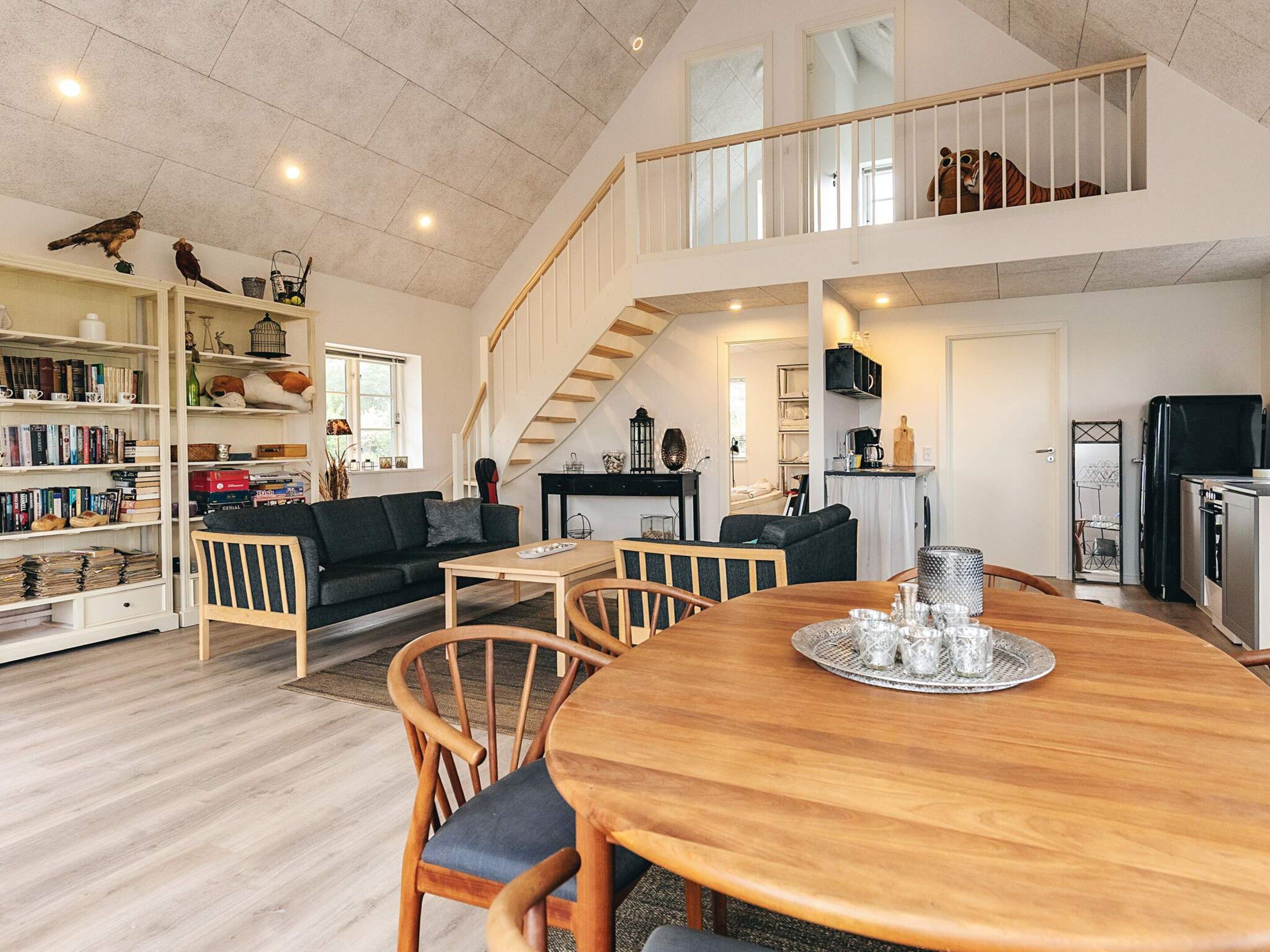 A spacious living area with a high ceiling, a staircase, a dining table with chairs, and a kitchen area.