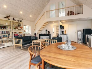 Amplia sala de estar con techo alto, una escalera, una mesa de comedor con sillas y una zona de cocina.