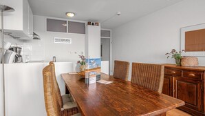 Een eethoek met een houten tafel en rieten stoelen, een keuken met witte kasten, en een rieten mand op de tafel.